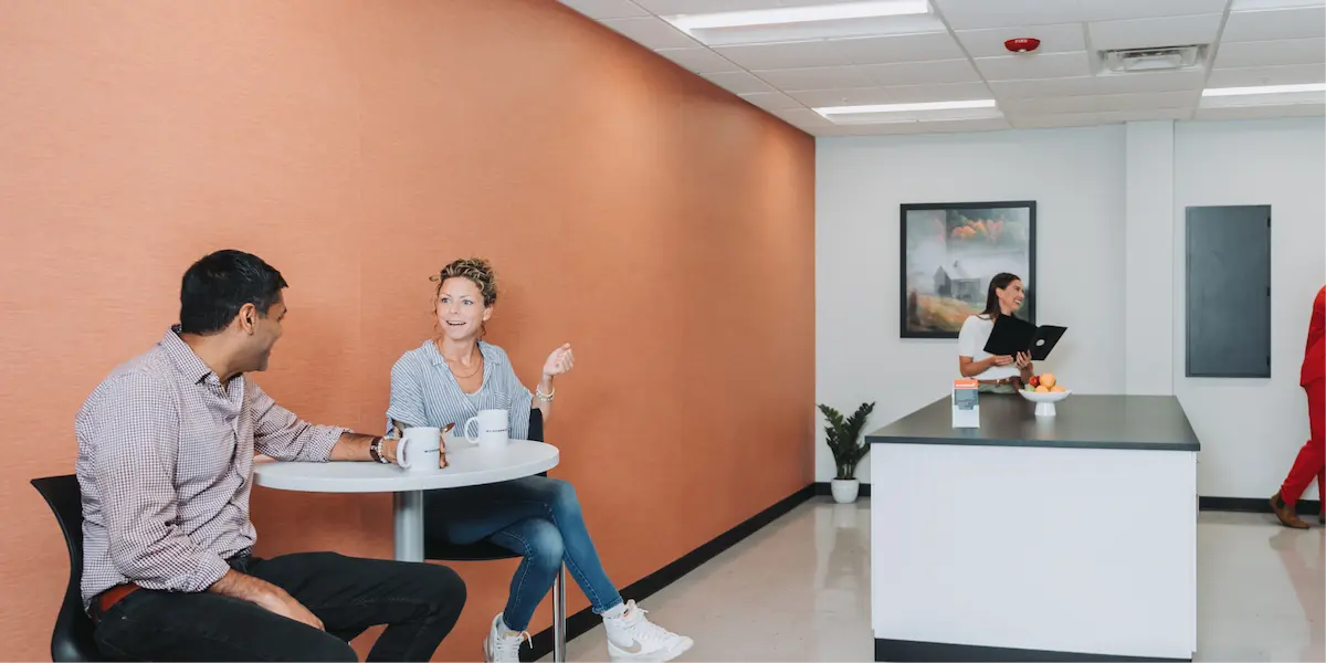 Two people sitting at a round table chatting over coffee in a modern break room, while a third person stands at a counter in the background.