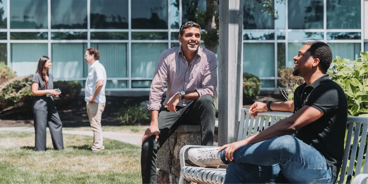 Two men smiling and talking outside near a bench, with another pair of colleagues conversing in the background.