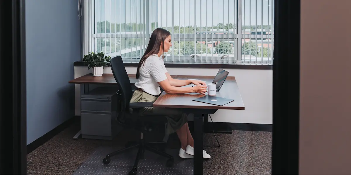 Woman working alone at a desk in a private office with a laptop and coffee mug, next to large windows with blinds.