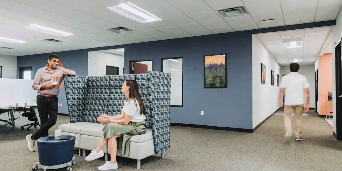 Man and woman having a discussion in a lounge area with high-backed seating, while another person walks down the hallway.