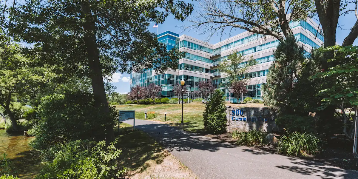 Exterior view of a modern glass office building surrounded by trees, with a pathway leading toward the entrance and a sign reading “500 Cummings Center.”