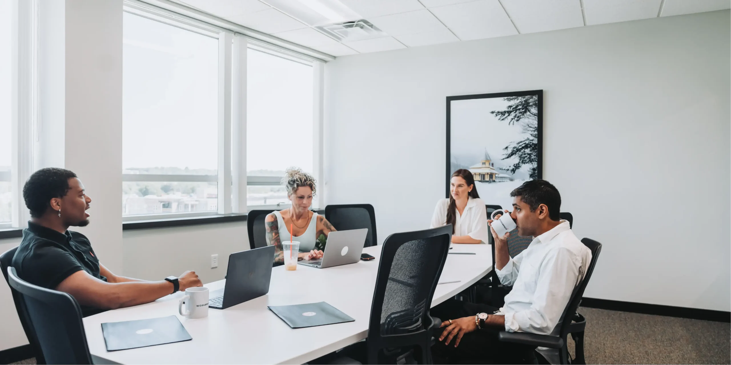 Four professionals seated around a white conference table in a bright meeting room, engaged in discussion, with laptops, notebooks, and coffee cups on the table.