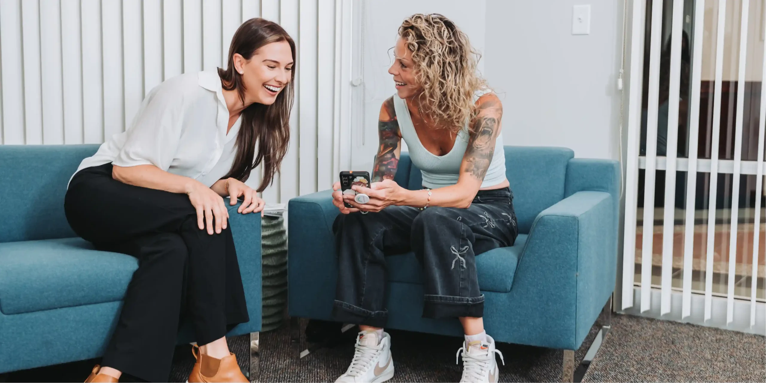 Two women sitting on teal armchairs in an office lounge, smiling and looking at a phone together.