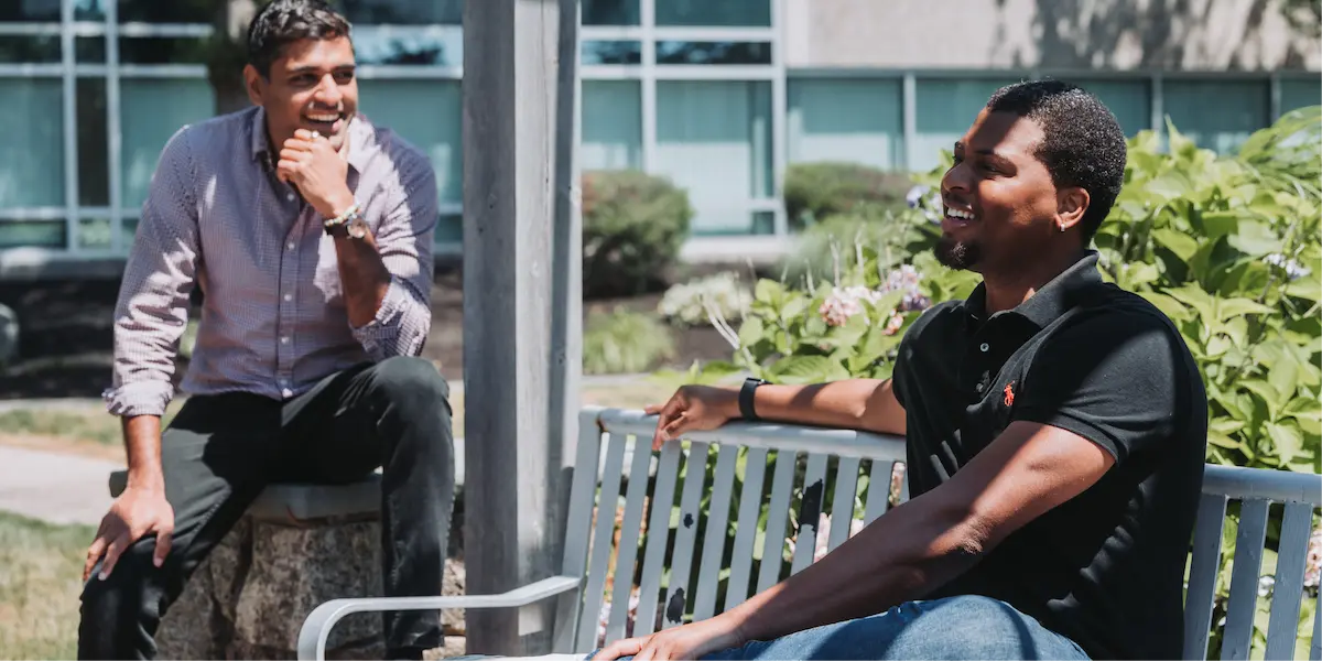 Two men seated outdoors in a landscaped area, talking and laughing in the sunshine.