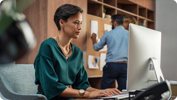 Coworking space operator working at desktop computer while a colleague updates a wall board in the background — representing workspace organization, systems, and operational efficiency